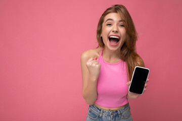 Beautiful joyful young blonde woman wearing pink top poising isolated on pink background with empty space holding in hand and showing mobile phone with empty display for mockup looking at camera and