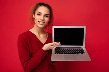 Naklejka premium Close-up portrait of smiling happy beautiful dark blond woman holding laptop computer looking at camera showing at netbook keyboard wearing red sweater isolated over red wall background