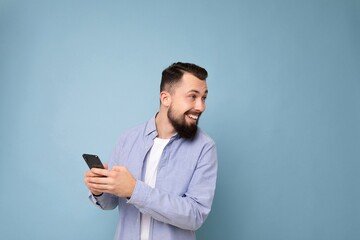 Photo shot of handsome positive good looking young man wearing casual stylish outfit poising isolated on background with empty space holding in hand and using mobile phone messaging sms looking behind