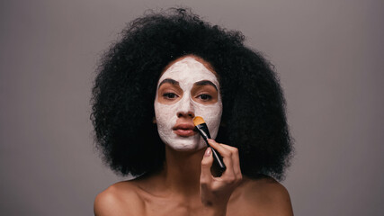Obraz premium curly african american woman with bare shoulders applying clay mask isolated on grey