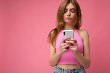 Photo shot of concentrated attractive positive good looking young woman wearing casual stylish outfit poising isolated on background with empty space holding in hand and using mobile phone messaging