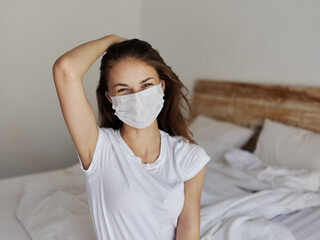 pretty woman in medical mask sits on the bed and touches her head with her hand