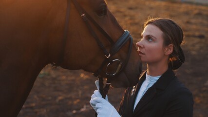 a young beautiful woman near the horse's head with gloved hands holding the reins and looking at the horse which touches her face. High-quality photo