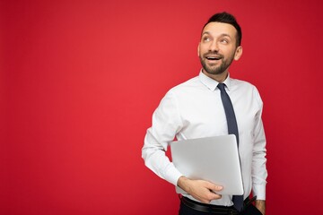 Handsome brunet man holding laptop computer looking up in white shirt and tie on isolated red background