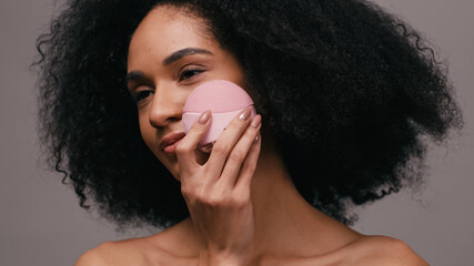 Naklejka premium curly african american woman using cleansing gadget on face isolated on grey