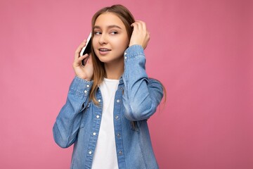 Photo of beautiful thoughtful young blonde woman wearing casual blue jean shirt isolated over pink background holding in hand and talking on mobile phone looking at camera and thinking