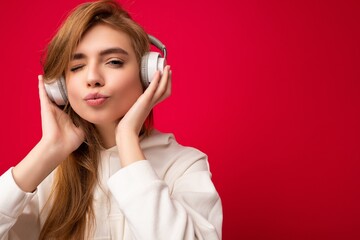 Closeup photo of attractive cute positive young blonde woman wearing white hoodie isolated over colourful background wearing white wireless bluetooth headphones listening to good music looking at