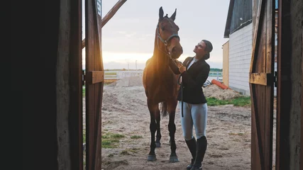 Foto op Plexiglas a young woman stands upright next to a horse she is holding by the halter in front of the entrance to the barn illuminated by natural light. High-quality photo © CameraCraft