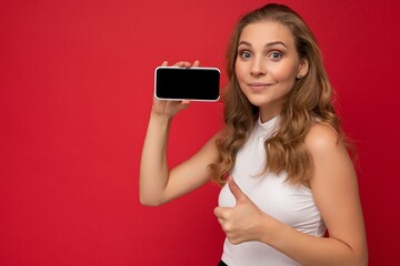 Portrait of happy beautiful young blonde woman wearing white t-shirt isolated on red background with copy space holding smartphone showing phone in hand with empty screen for mockup looking at camera