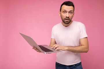 Handsome surprised and amazed man holding laptop computer looking at camera and texting in t-shirt on isolated pink background