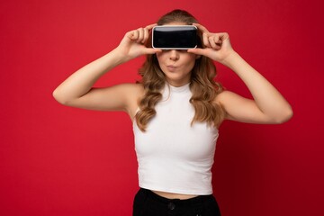 funny young blonde woman wearing white t-shirt isolated on red background with copy space holding smartphone showing phone in hand with empty screen for cutout