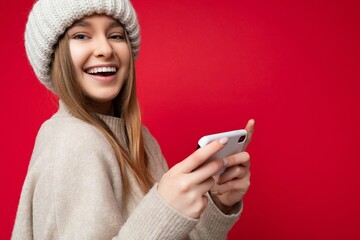 Closeup portrait of sexy smiling happy Beautiful young blonde woman wearing casual beige sweater and beige hat isolated over red background holding in hand and using mobile phone communicating online