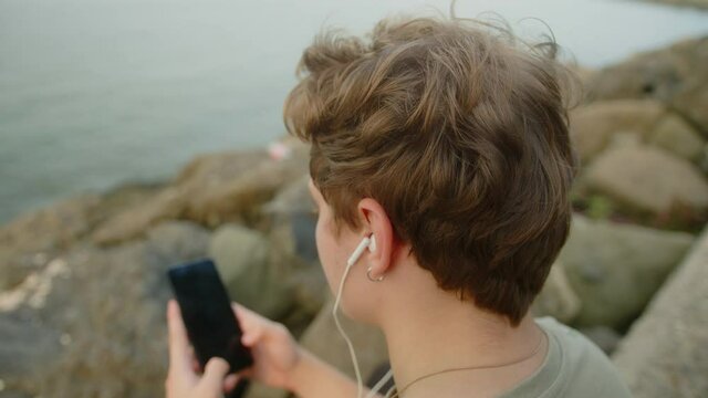 Young blond boy with earring sitting on beach looking his phone and listening to music in the fresh air alone.