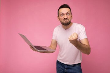 Handsome angry and wrathful man holding laptop computer showing fist looking at camera in t-shirt on isolated pink background