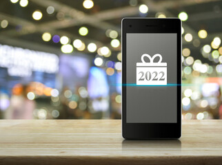 Gift box happy new year 2022 flat icon on modern smart mobile phone screen on wooden table over blur light and shadow of shopping mall, Business happy new year 2022 shop online concept