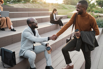 Horizontal high angle shot of two African American men wearing fashionable elegant clothes greeting each other with handshake outdoors © AnnaStills
