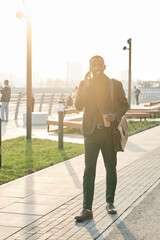 Vertical full body shot of handsome young African American man wearing fashionable outfit walking along street talking on phone and drinking fresh coffee after workday