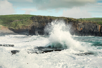 Powerful waves crushing against Cliffs and rough stone coastline of West coast of Ireland. Doolin area. County Clare. Ocean power and rugged Irish coastline. Green fields in the background