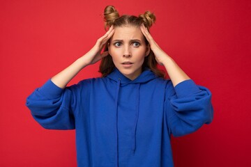 Photo of emotional shocked young pretty blonde woman with two horns with sincere emotions wearing hipster bright blue hoodie isolated over red background with copy space