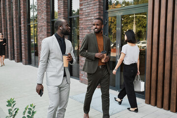 Horizontal medium long shot of two handsome Black men wearing fashionable outfits holding cups with coffee walking and talking during break time