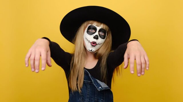 Scary bright alive fun girl child with Halloween makeup mask wears black hat, hands raised, spooky looking at camera, isolated over yellow color background wall in studio. Party holiday concept