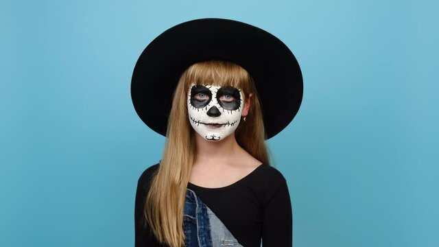 Portrait of spooky child girl with Halloween makeup mask wears big black hat looking at camera you blow send air kiss flirting, posing isolated over blue color background in studio. Holiday concept