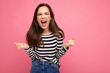 Portrait of young positive happy beautiful brunette woman with sincere emotions wearing casual striped pullover isolated on pink background with empty space and celebrating victory shouting yes