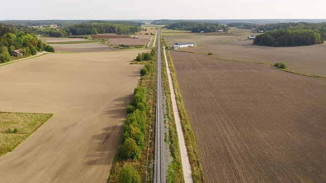 High altitude tracking aerial drone view of high speed passenger train approaching on long straight railroad with fields and forests on both sides. Passing under road bridge.