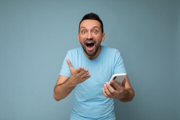 Amazed emotional handsome young bearded man wearing everyday blue t-shirt isolated over blue background holding and using mobile phone communication online on the internet looking at camera