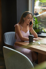 beautiful young asian woman sitting at coffee shop table surrounded by plants, working on laptop. beautiful lady calling by smartphone. modern online education, distant work, remote job, freelance
