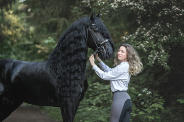 Beautiful young lady posing with a purebred friasian horse in the forest. - 459062650