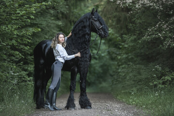 Beautiful young lady posing with a purebred friasian horse in the forest. - 459062653