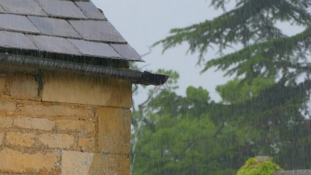 Overflowing water during heavy rainfall, at the end of a piece of roof guttering, running down the wall of a centuries old stone house, with large trees swaying behind in the wind.