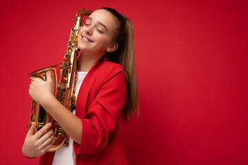 Fototapeta na wymiar Fascinating cute positive smiling brunette little female teenager wearing trendy red jacket standing isolated over red background wall holding saxophone and dreaming with close eyes