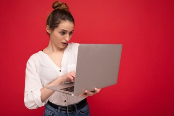 Side profile of Beautiful amazed brunet young woman holding netbook computer looking down with open mouth wearing white shirt typing on keyboard isolated on red background