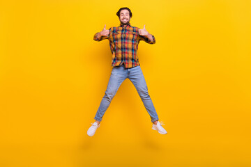 Full length photo of cool brunet millennial guy jump show thumb up wear shirt jeans sneakers isolated on yellow background
