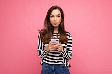 Beautiful young brunette woman using mobile phone communicating via texting message wearing sweater isolated on wall background looking at camera