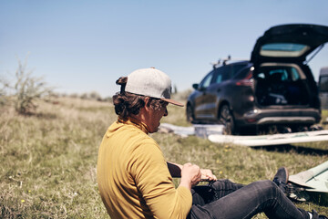 Windsurfer and camper packing and unpacking from a car's roof rack in nature.