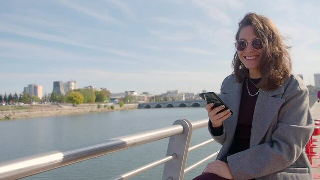 Woman typing text message on her smartphone outdoor while sitting on the city river coast.