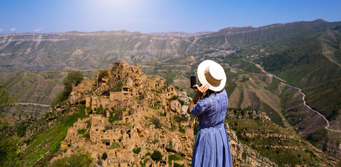 Fototapeta na wymiar Female tourist high in mountains of Dagestan admires the breathtaking panoramic view of nature and the ancient abandoned city of Gamsutl. Takes photos attraction and ruins, selfies on a smartphone