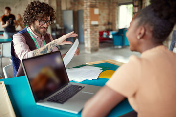 caucasian beardy guy with glasses and tattoo handing over a document to his afro-american female colleague