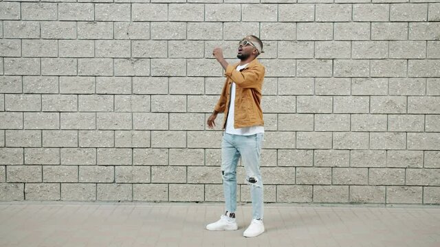 Funny Afro-American man is walking with robot moves drinking from imaginary bottle outdoors on brick wall background. People and art concept.