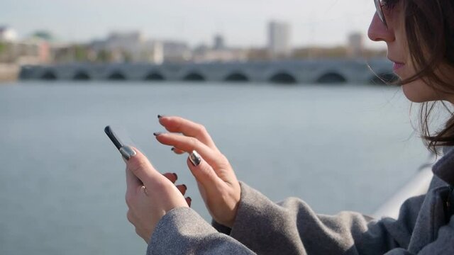 Close-up view of a woman typing text message on her smartphone outdoor while sitting on the city river coast.
