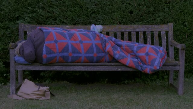 An unidentifiable person wearing winter clothing, having just got into a sleeping bag, lying down at twilight for a cold night on a wooden park bench, with some belongings nearby.