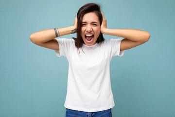 Fototapeta premium Portrait of emotional young beautiful brunette female person with sincere emotions wearing casual white t-shirt for mockup isolated on blue background with empty space and covering ears with hands and
