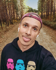 sporty man in a sweatshirt, baseball cap and headphones in the woods. Funny selfie portrait.