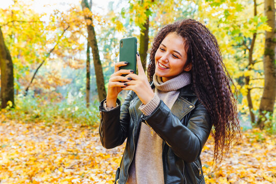Beautiful afro-haired woman walk in autumn park at sunny warm day. Portrait of woman outdoor making photos with smartphone