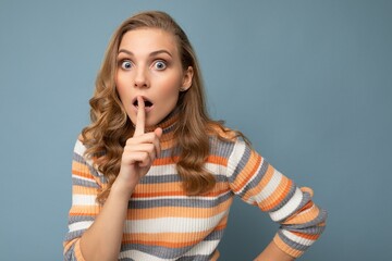 Shot of young amazed shocked blonde wavy-haired woman with sincere emotions wearing striped pullover isolated on blue background with copy space and showing shh gesture
