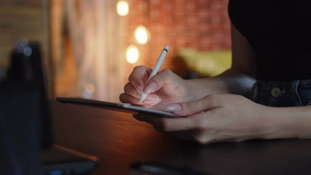 Woman artist draws on graphic tablet while sitting at table using stylus, side view, closeup. Female designer works on digital tablet using digital pen
