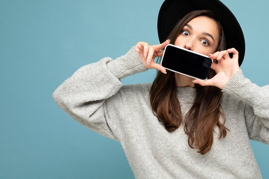 Closeup Photo of Beautiful positive woman person wearing black hat and grey sweater holding mobilephone showing smartphone isolated on background looking at camera